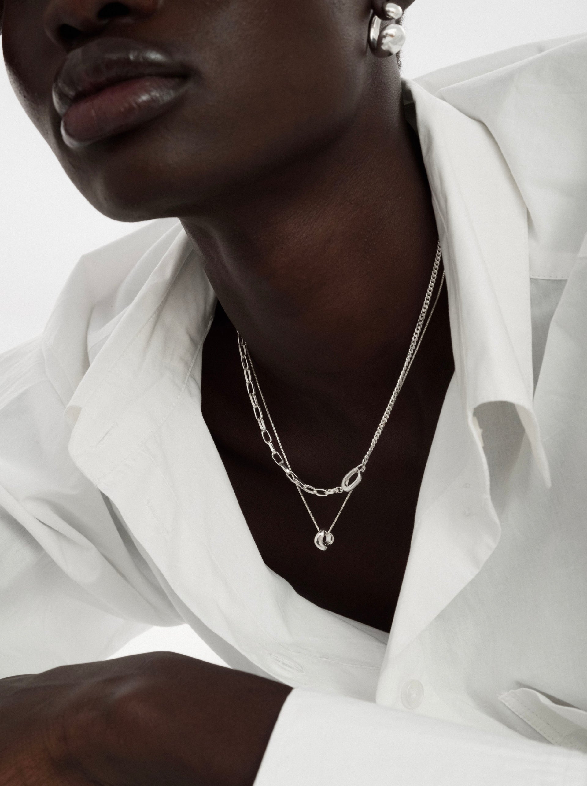 A close-up of a model wearing the Bolded Wavy Necklace in silver, featuring a fine chain and a small wavy pendant, layered with another silver chain necklace and paired with a chunky silver earring.