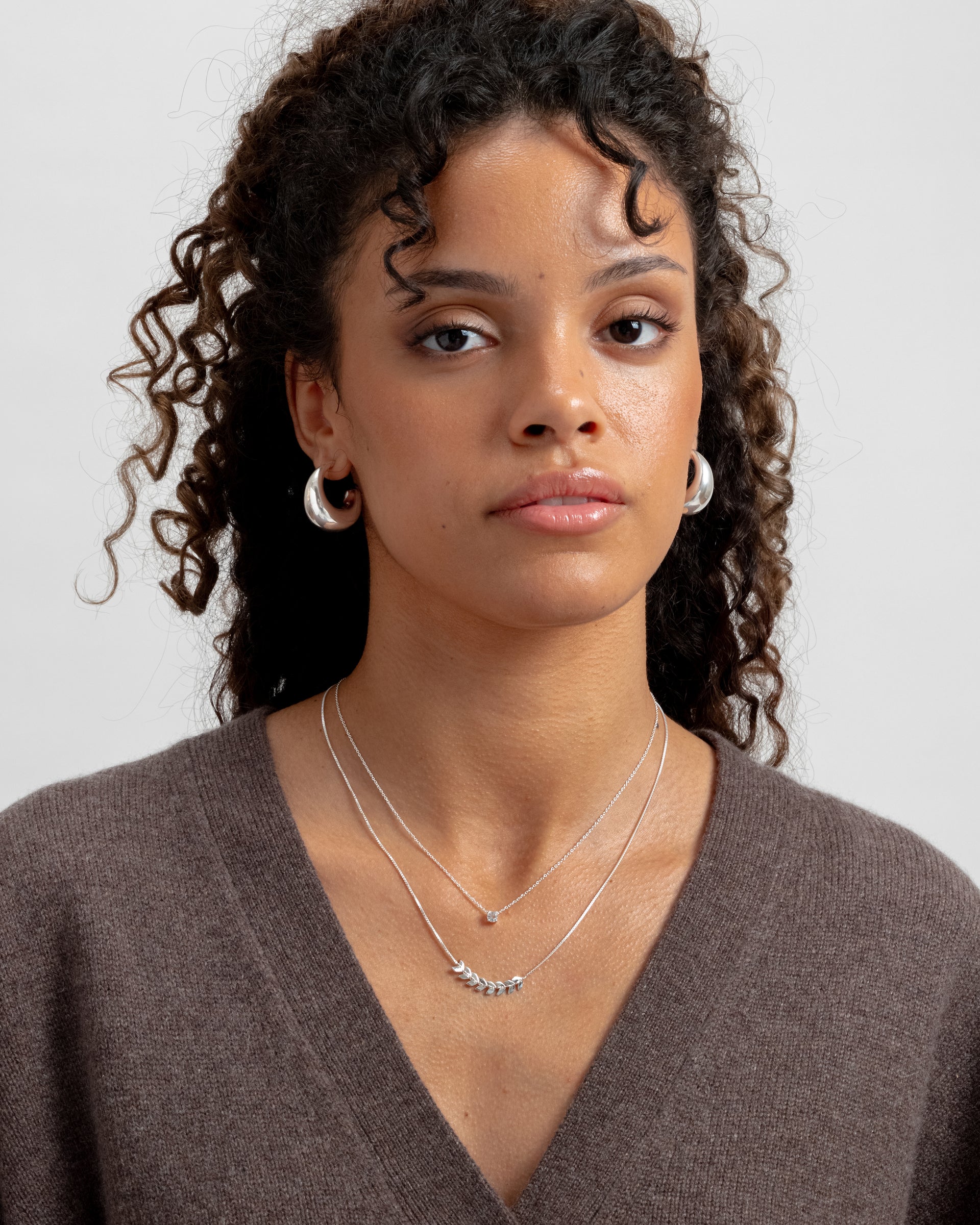 A model shot of a woman wearing the Minimalistica Solo Nova Necklace in silver, a delicate piece with a single white topaz, layered with a second silver necklace featuring a leaf motif, and paired with thick silver hoop earrings.