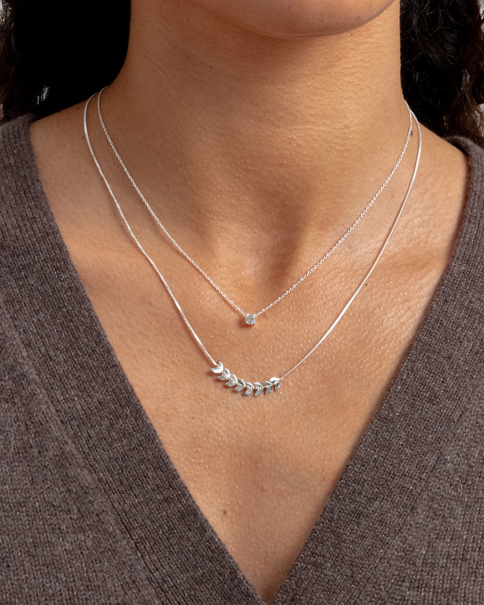 A close-up of a model wearing the Minimalistica Solo Nova Necklace in silver, layered with another silver necklace featuring a central leaf-like design.