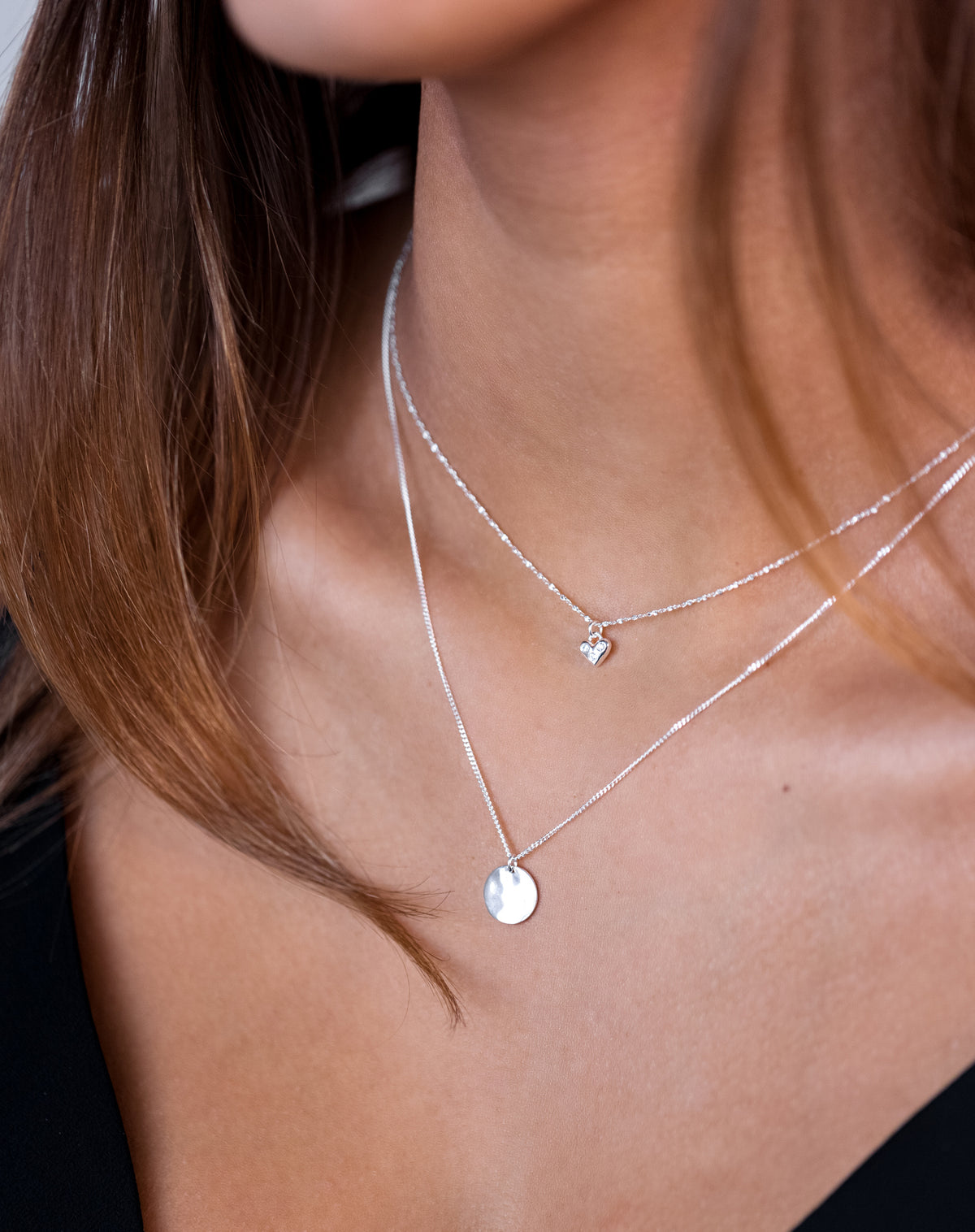 A close-up shot of a model wearing the Forever Necklace in silver, featuring a delicate heart pendant with sparkling stones, layered with a longer silver chain necklace with a round pendant.