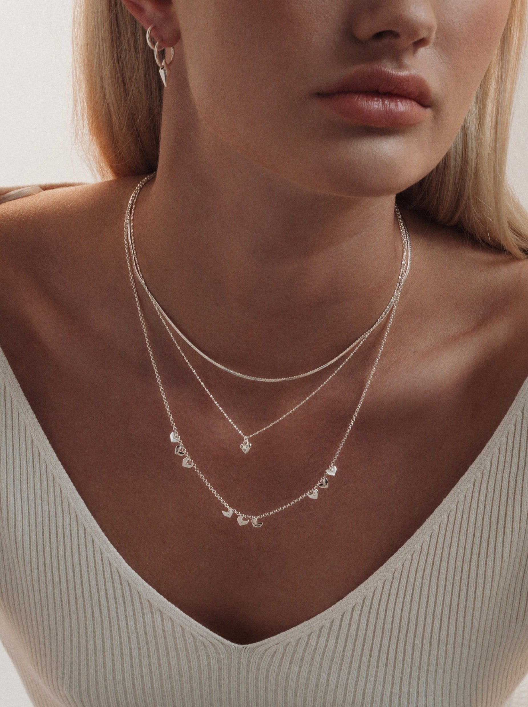 A model wearing the silver Forever Necklace with its sparkling heart pendant, layered with a silver snake chain, a longer necklace with small heart charms, and small silver spike hoop earrings.