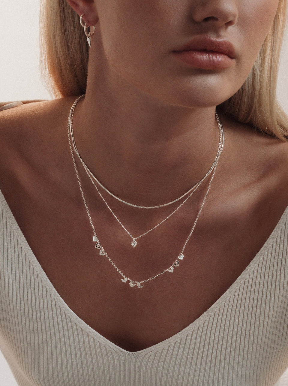 A model wearing the silver Forever Necklace with its sparkling heart pendant, layered with a silver snake chain, a longer necklace with small heart charms, and small silver spike hoop earrings.