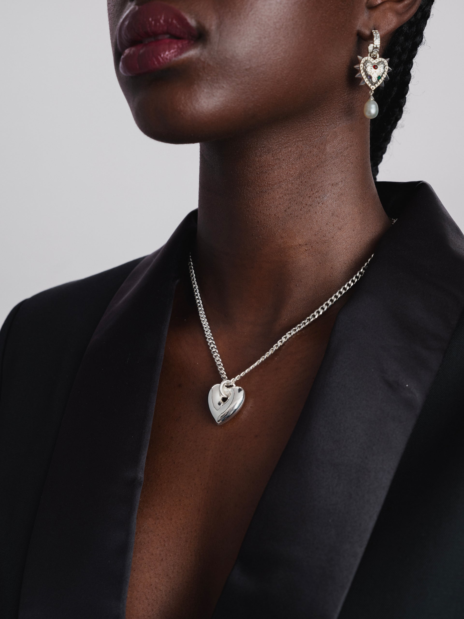 A close-up of a model wearing the Fat Heart Necklace in silver, featuring a chunky chain and a large heart pendant, along with a silver earring with a spiky heart design and a dangling pearl.