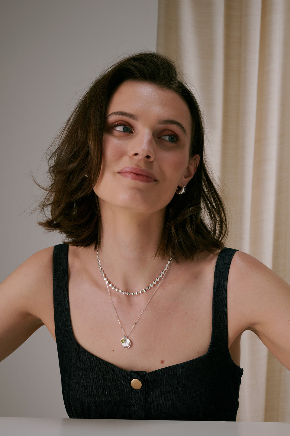 A silver necklace with a chain of small linked hearts, layered with a longer silver pendant necklace and paired with small silver hoop earrings.