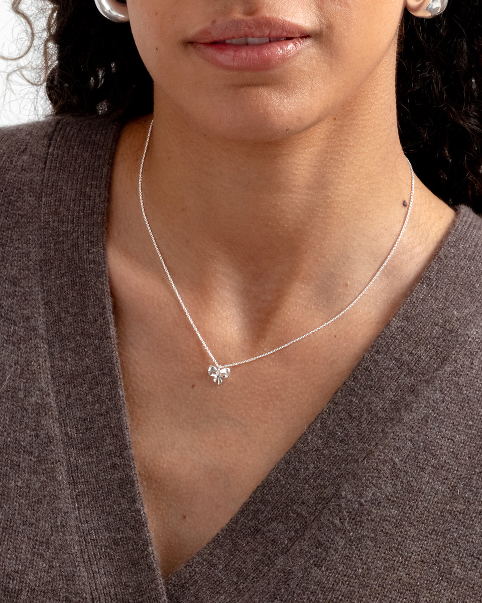 A close-up of a model wearing the Rosie Mini Necklace in silver, featuring a structured, sparkling chain and a small bow pendant, complemented by chunky silver hoop earrings.