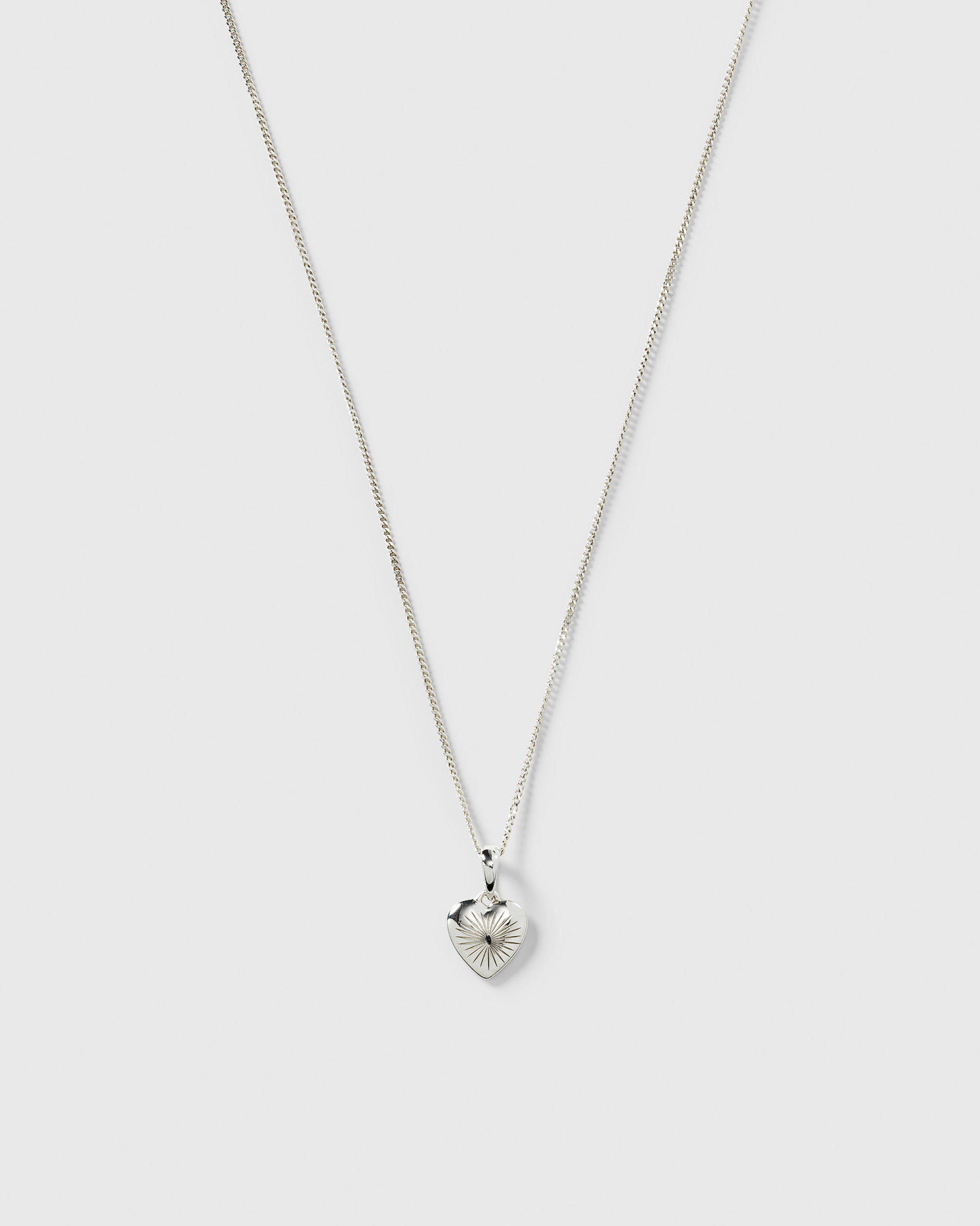 A product shot of the Sunburst Heart Necklace in silver, featuring a fine chain and a heart-shaped pendant with an engraved sunburst pattern.