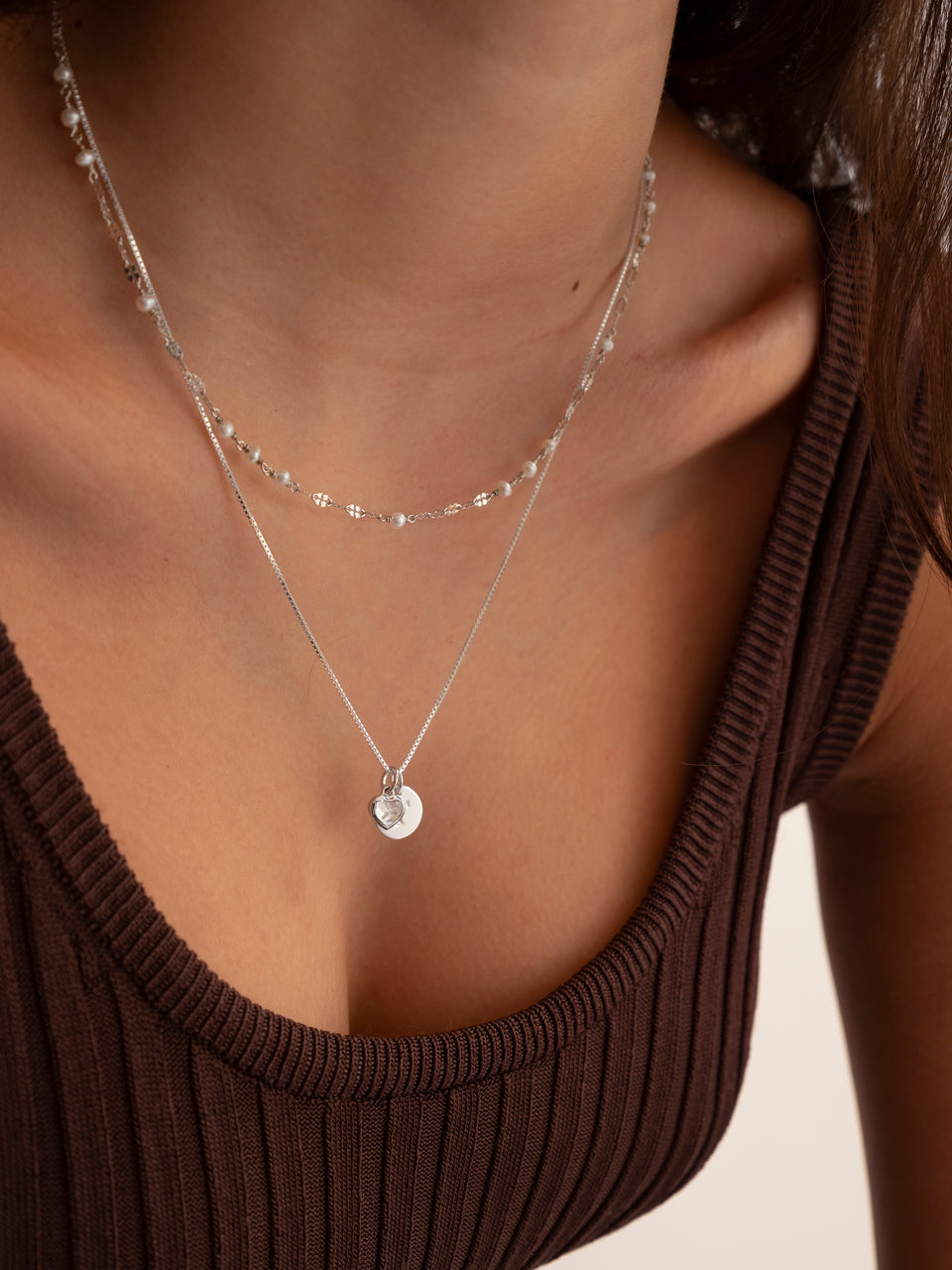 A close-up of a model wearing two layered silver necklaces. The longer necklace features the Beloved Heart Pendant with a crystal heart and a flat silver disc on a box chain. The shorter necklace is a delicate chain with small pearls.