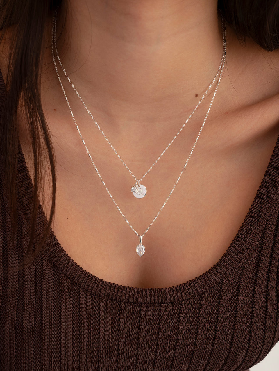 A model wears the Katie Necklace in silver, featuring a unique chain and a drop-shaped cubic zirconia pendant, layered with a second silver necklace that has a small, round engraved charm.
