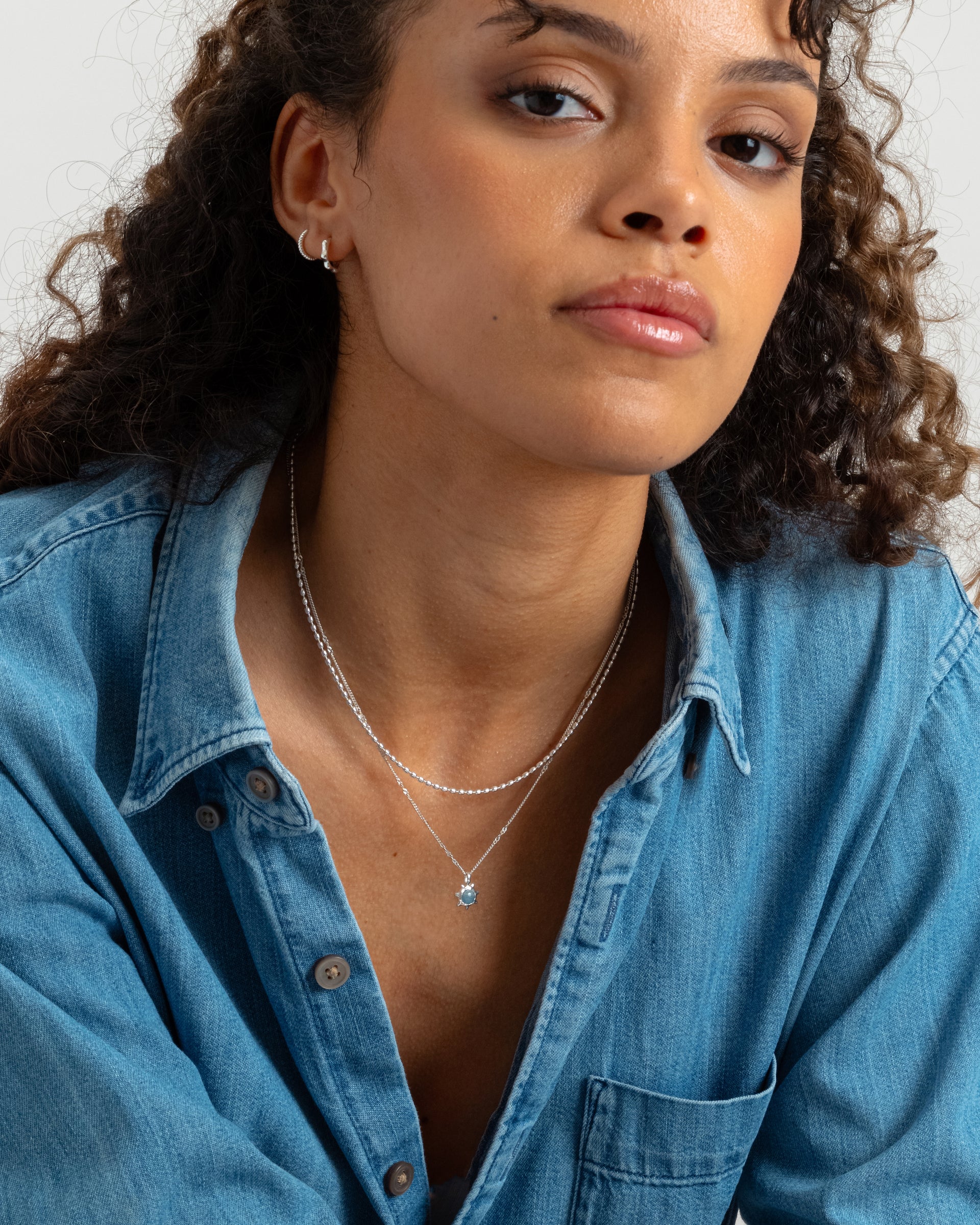 A close-up shot of a model wearing the Florentina Necklace in silver, featuring a flower-shaped pendant with a light blue aquamarine stone. The necklace is layered with another silver chain, and she is also wearing two small silver hoops in her ear.