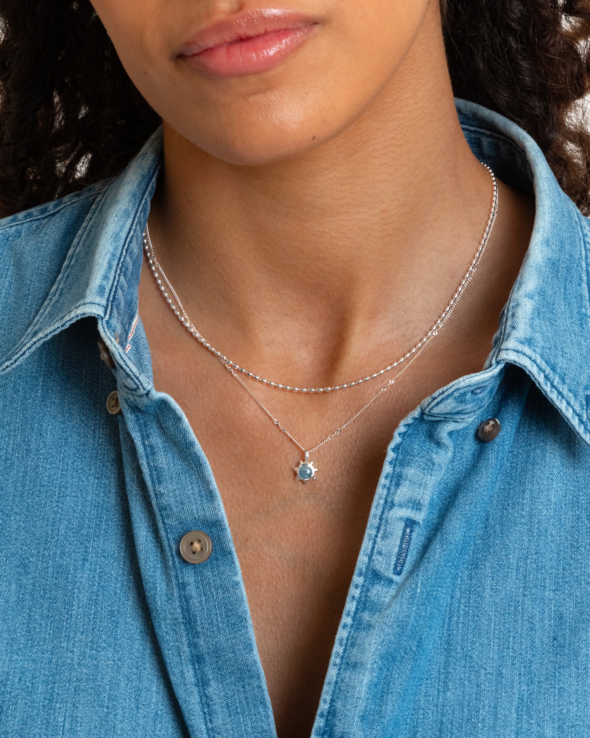 A close-up of a model wearing the Florentina Necklace in silver, featuring a flower-shaped pendant with an aquamarine stone, layered with a shorter silver beaded chain necklace.