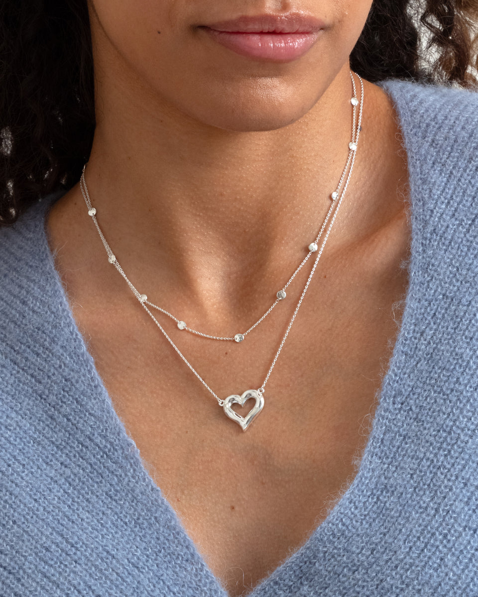 A model wears the Puff Heart Necklace in silver, featuring a chunky open-heart pendant, layered with a delicate silver chain necklace adorned with small, round hammered circles.