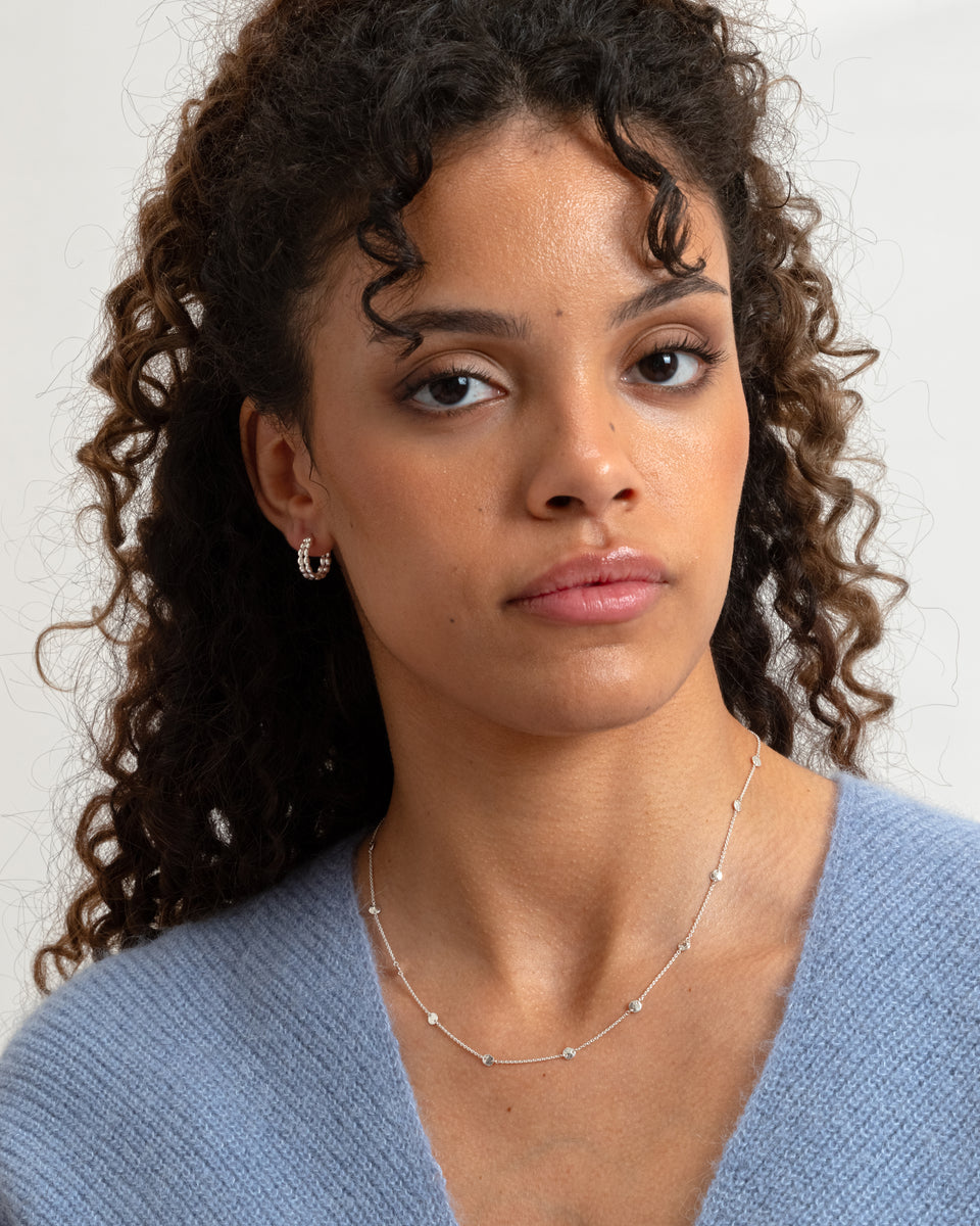 A model wears the Solea Necklace in silver, featuring a delicate chain with small, hammered round circles, paired with a textured silver hoop earring.