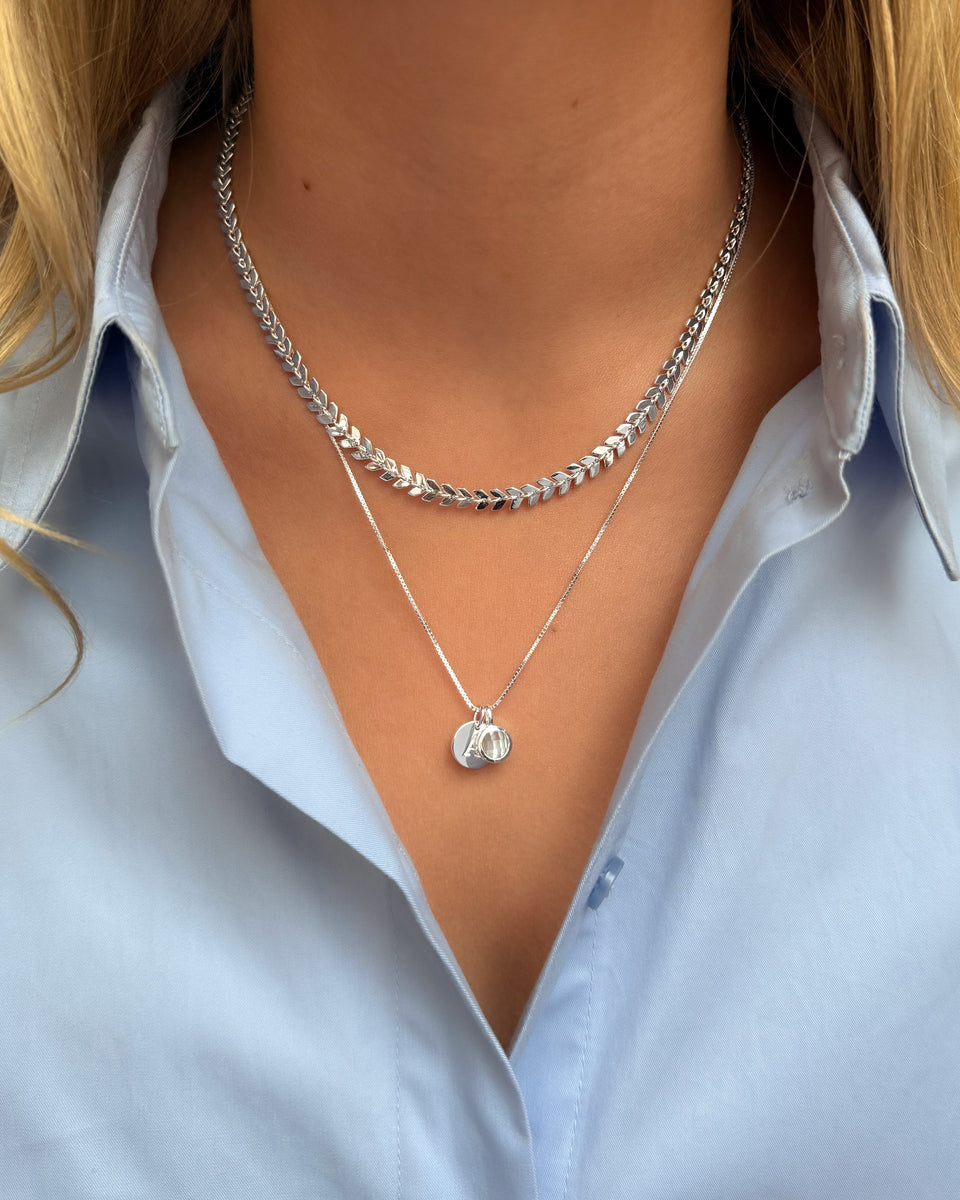 A necklace with a delicate leaf design, layered with a longer necklace featuring a March birthstone pendant set with light blue aquamarine, accented by a round letter charm.
