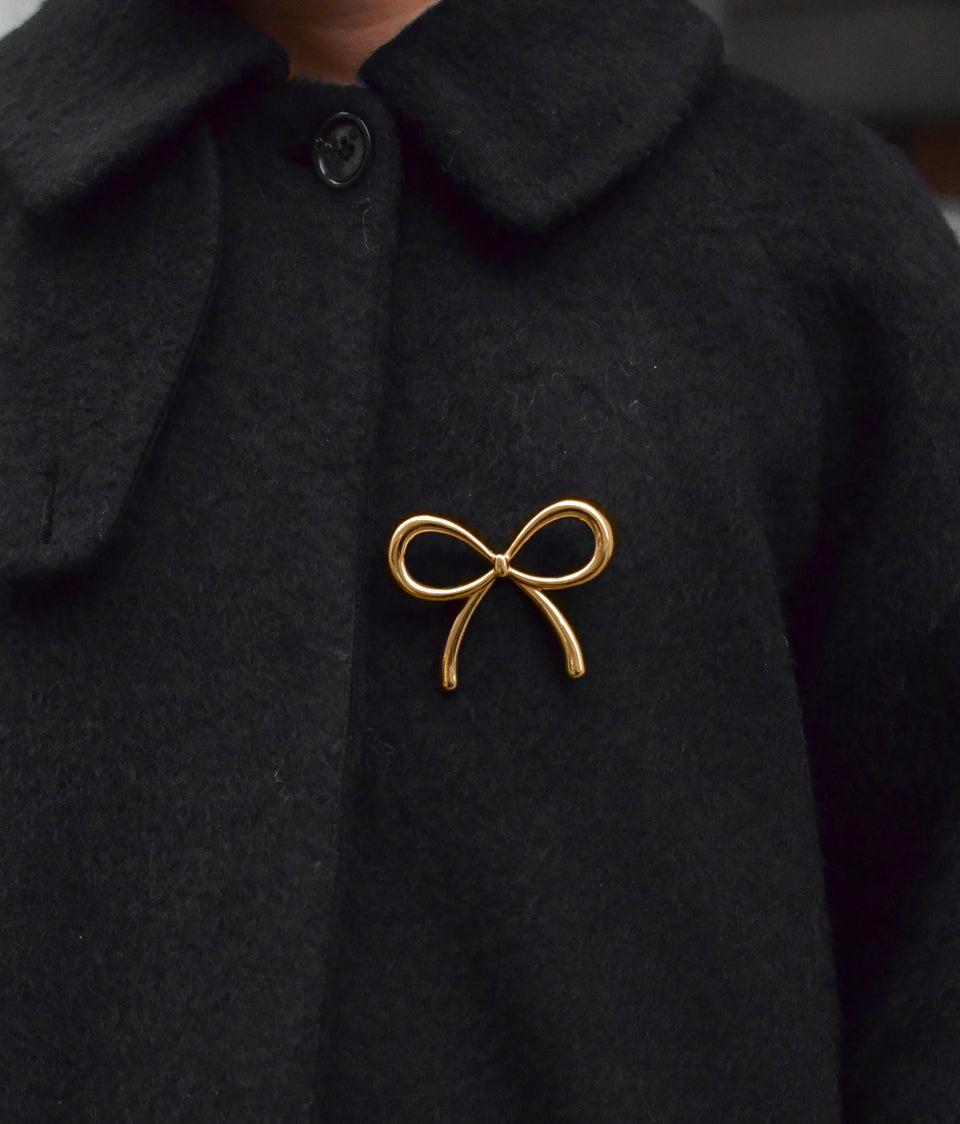 A close-up of the Ana Brooch in gold, featuring a delicate bow design, pinned onto a black coat.