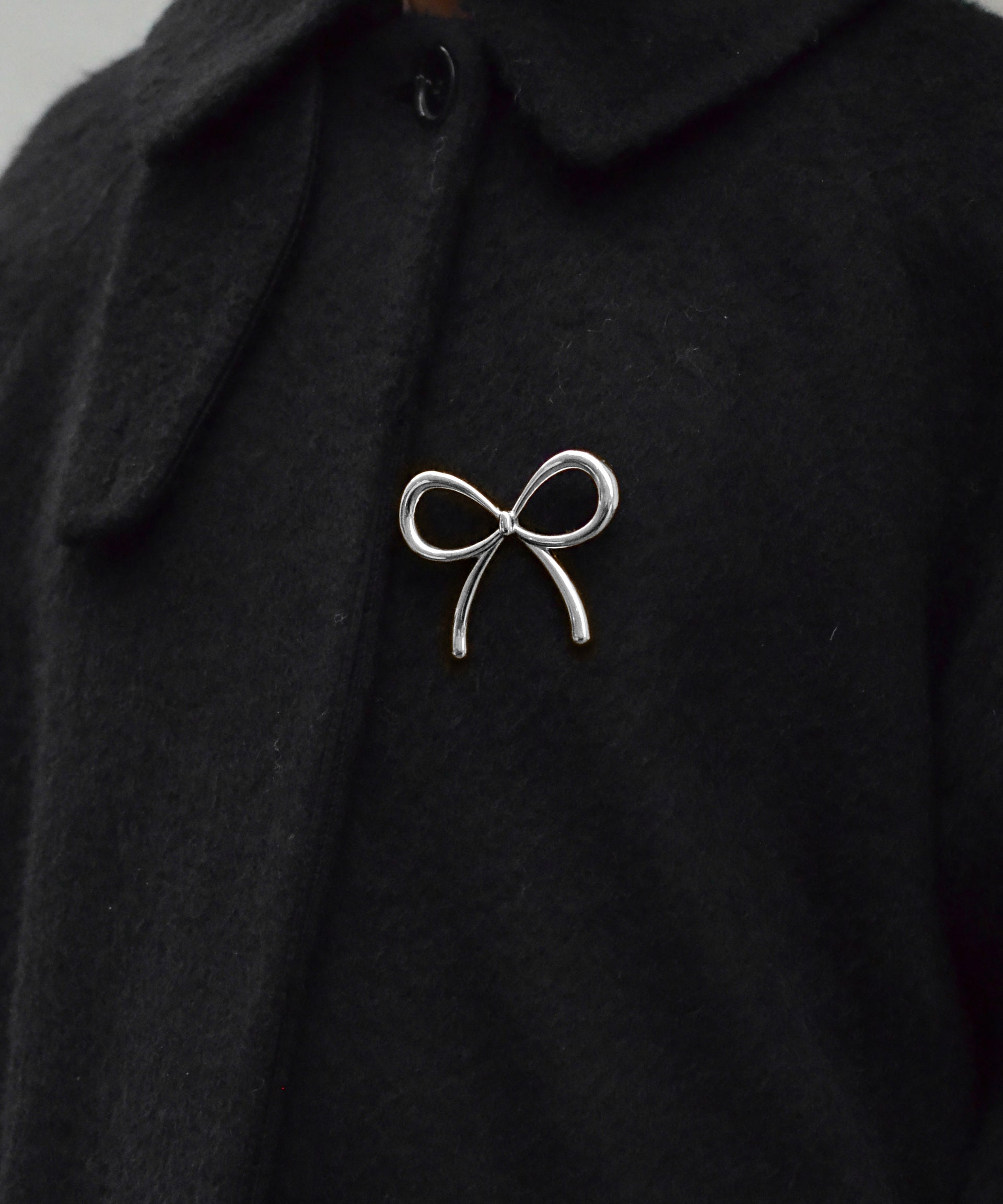 A model wears the Ana Brooch in silver, a playful bow-shaped design, pinned to the front of a black coat.