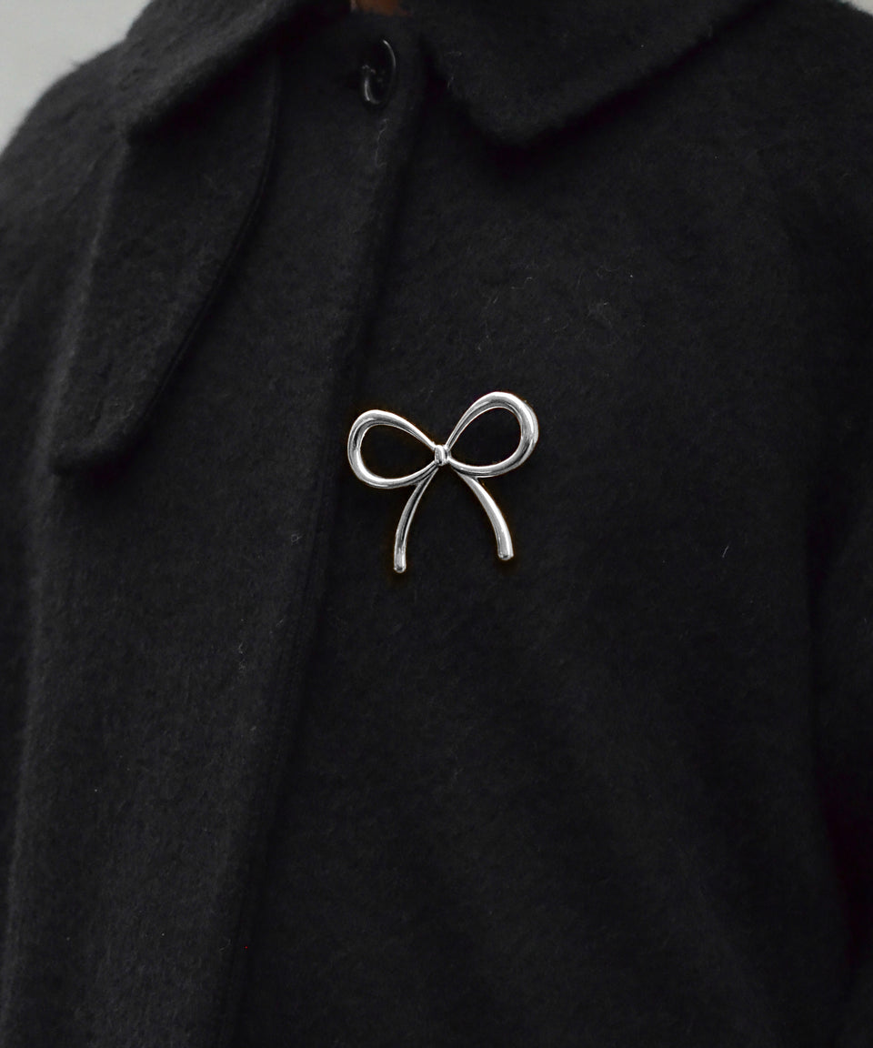 A model wears the Ana Brooch in silver, a playful bow-shaped design, pinned to the front of a black coat.