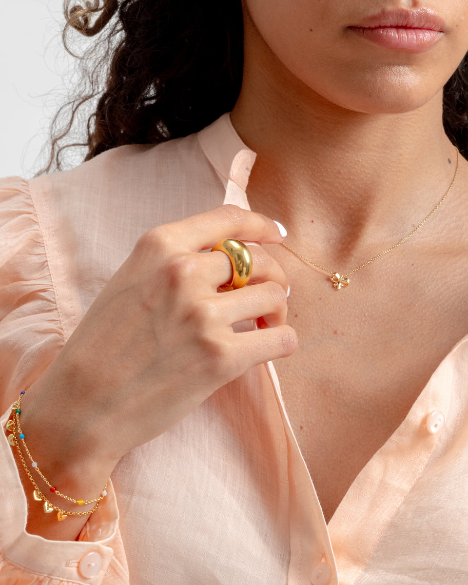 A close-up of a model wearing the Bolded Big Ring in gold, styled with a delicate gold necklace with a clover pendant and a layered gold bracelet with colorful beads and heart charms.