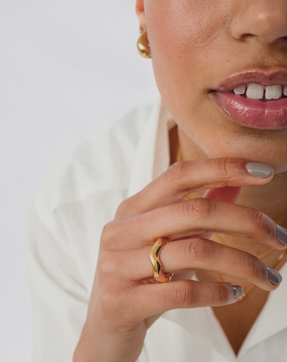 A close-up shot of a model wearing the Bolded Wavy Ring in gold, along with small gold earrings and a delicate gold chain necklace.