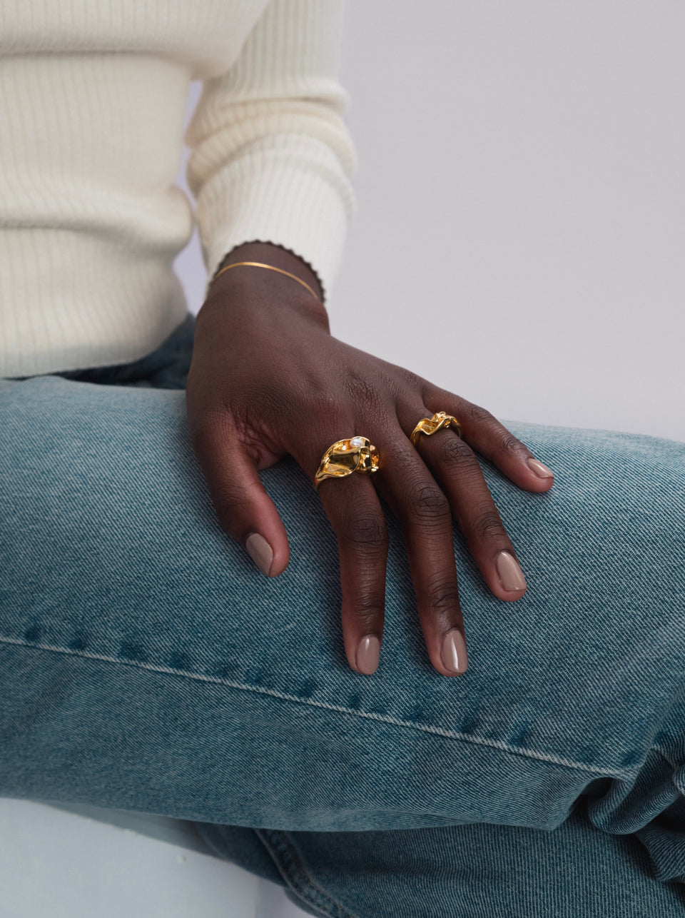 A close-up of a model wearing the Moulded Big Ring Gold, an irregularly shaped ring with a unique freshwater pearl, paired with a smaller wavy gold ring and a delicate gold bracelet.