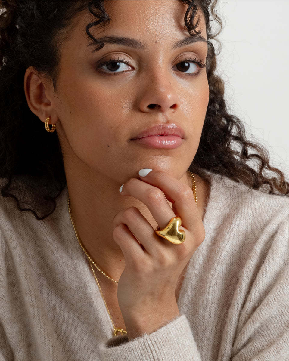 A close-up of a model wearing the Bolded Heart Ring in gold, a statement heart-shaped ring, complemented by small gold hoops and a gold necklace.