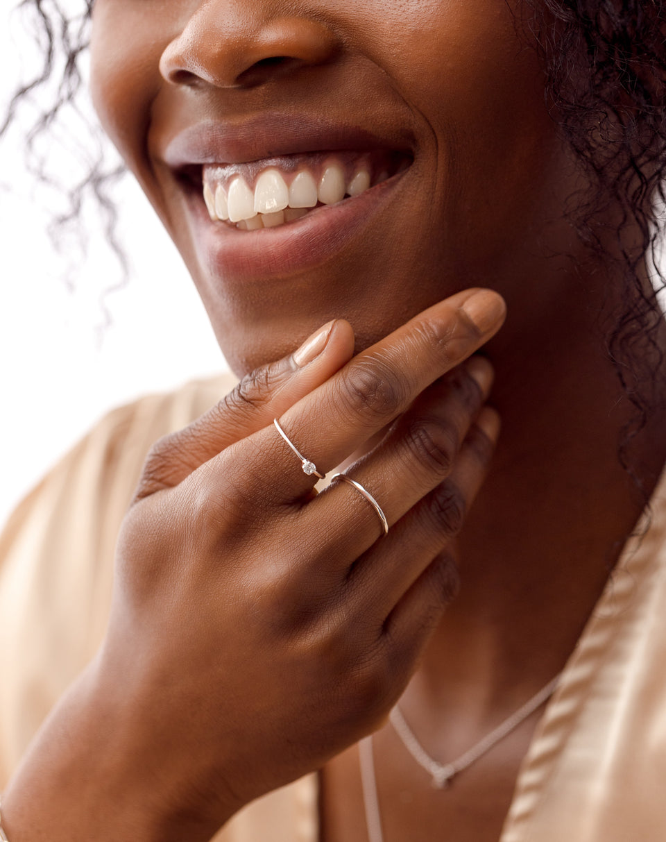 A close-up shot of a smiling model wearing the Tiny Princess Ring in silver, featuring a delicate band with a single crystal, alongside a plain silver band on her middle finger and a matching thin necklace.