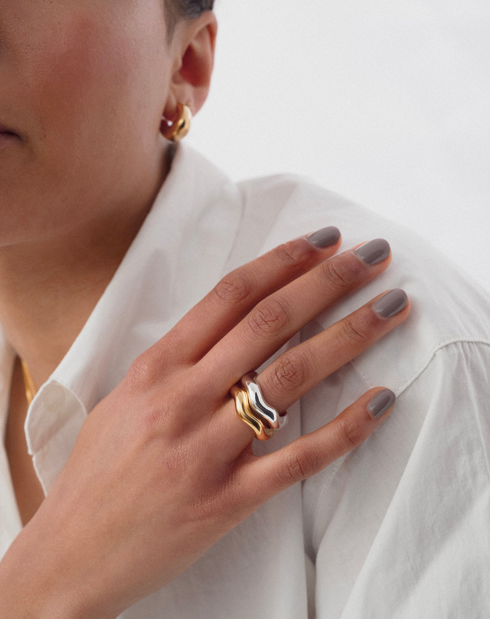 A close-up shot of a model wearing the Bolded Wavy Ring in silver stacked with the matching gold version, complemented by a small gold hoop earring.