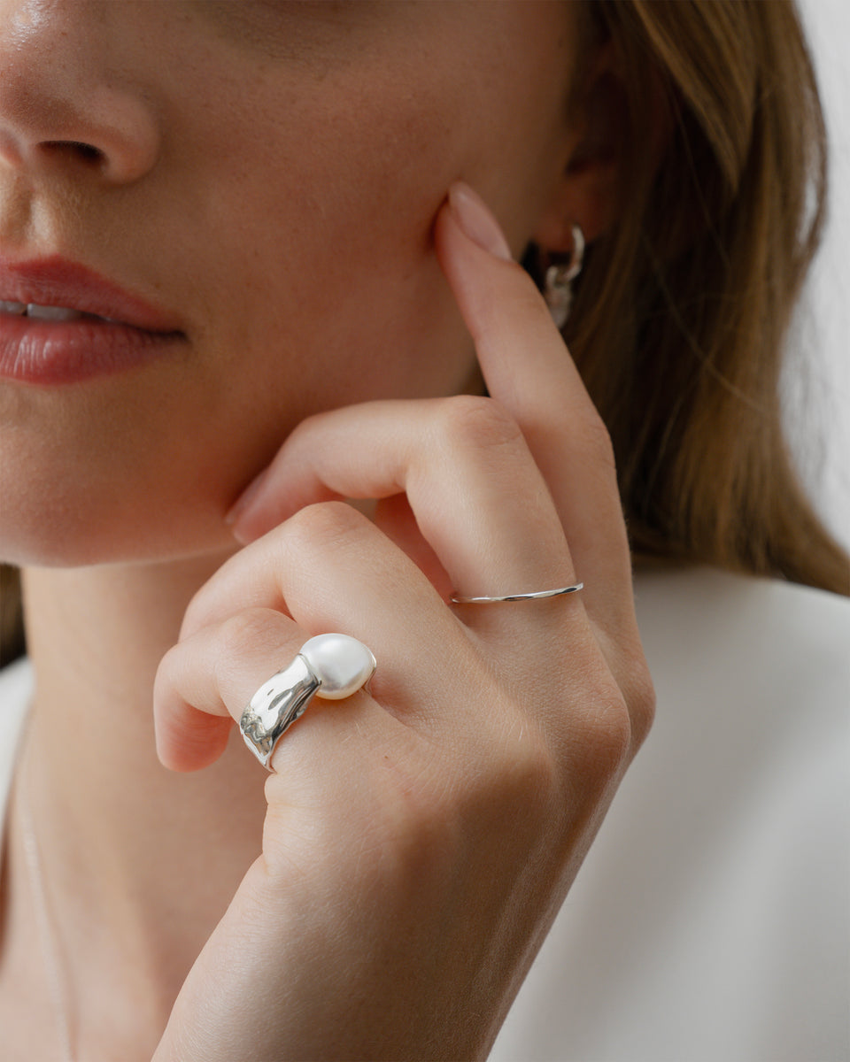 A close-up of a model wearing the Paxton Ring, a hammered silver ring with a large baroque pearl. She also wears a thin silver band on her index finger and a small silver hoop earring with a pendant.