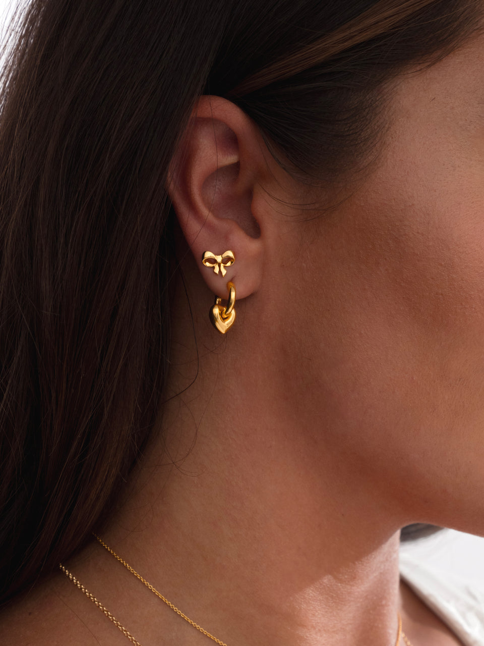 A close-up of a model's ear showing the Rosie Mini Studs in gold, shaped like a bow, styled with the gold LaLaLove Fat Heart Earrings and a fine gold chain necklace.