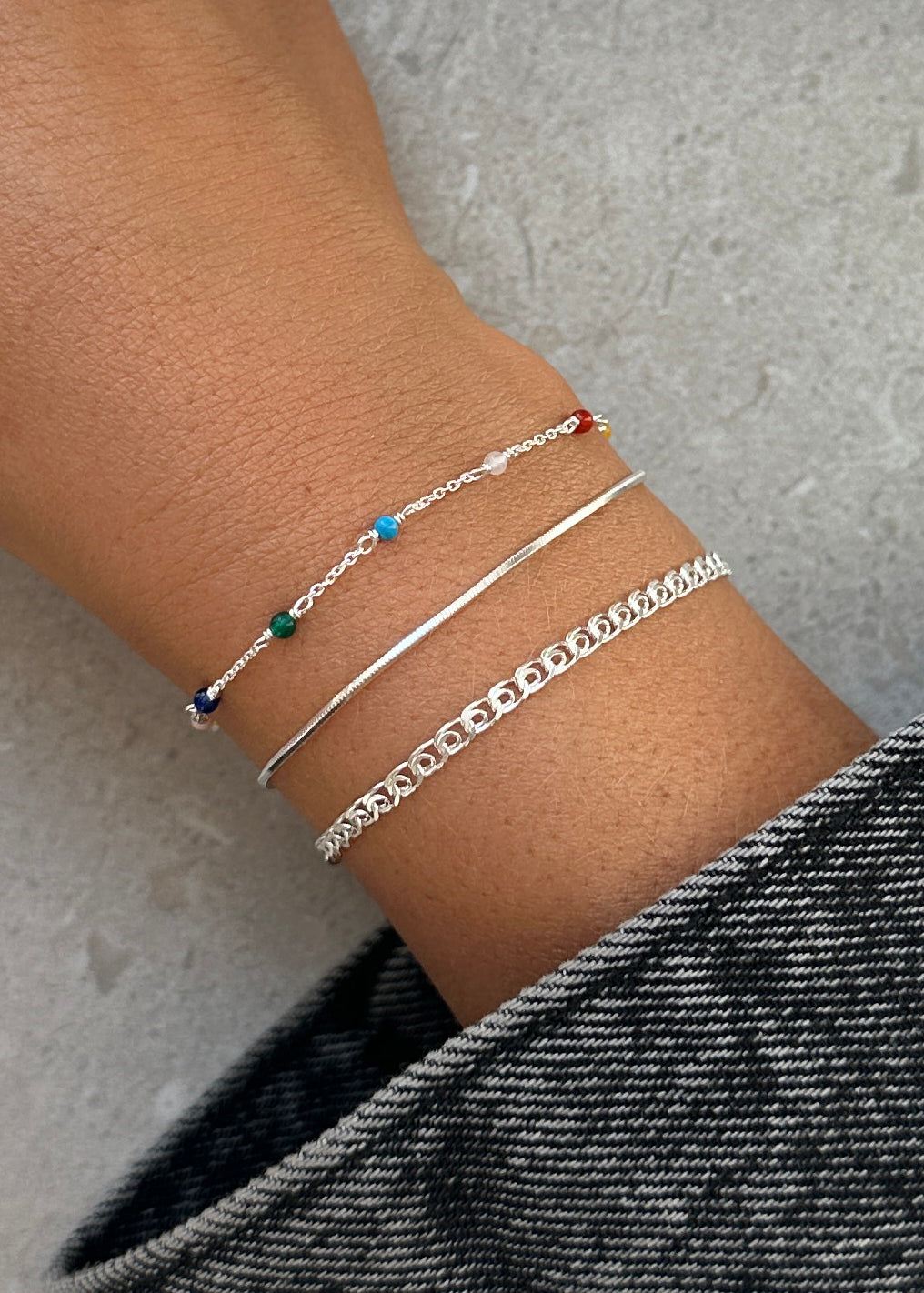 A model's wrist styled with a stack of three Syster P silver bracelets: the Santa Monica Bracelet with colorful gemstones, the simple Sheer Mirror chain bracelet, and the Sheer Love linked chain bracelet.