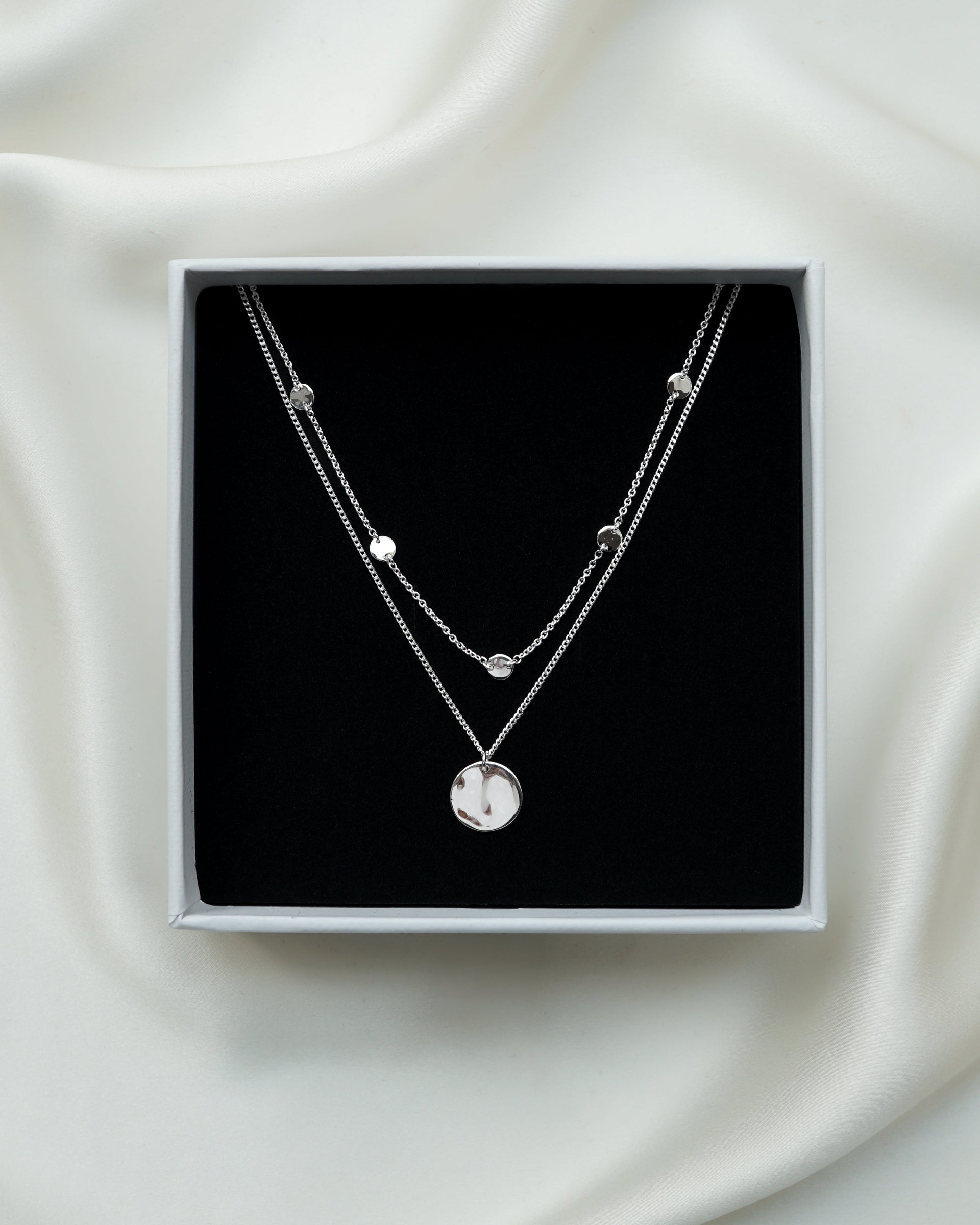 A close-up of a jewelry box with the Solea Necklace Silver, which has a delicate chain adorned with small hammered silver circles, layered with another silver necklace featuring a round hammered pendant.