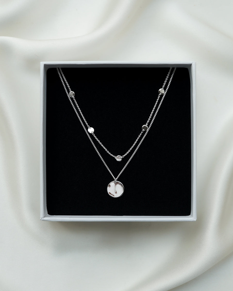 A close-up of a jewelry box with the Solea Necklace Silver, which has a delicate chain adorned with small hammered silver circles, layered with another silver necklace featuring a round hammered pendant.
