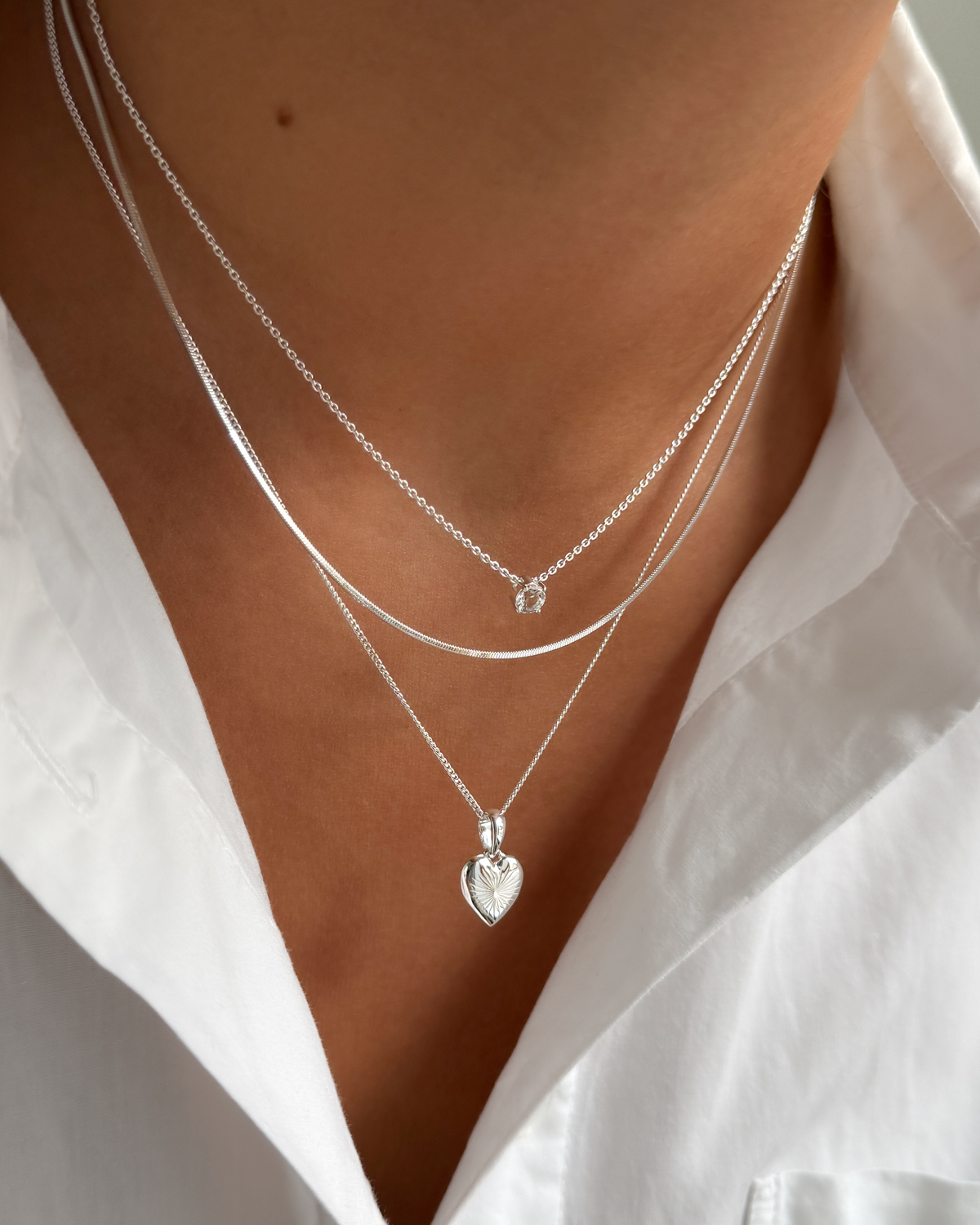 A close-up of a model wearing three layered silver necklaces: the top Minimalistica Solo Nova Necklace with a white topaz stone, the middle Sheer Mirror Necklace, and the bottom Sunburst Heart Necklace.
