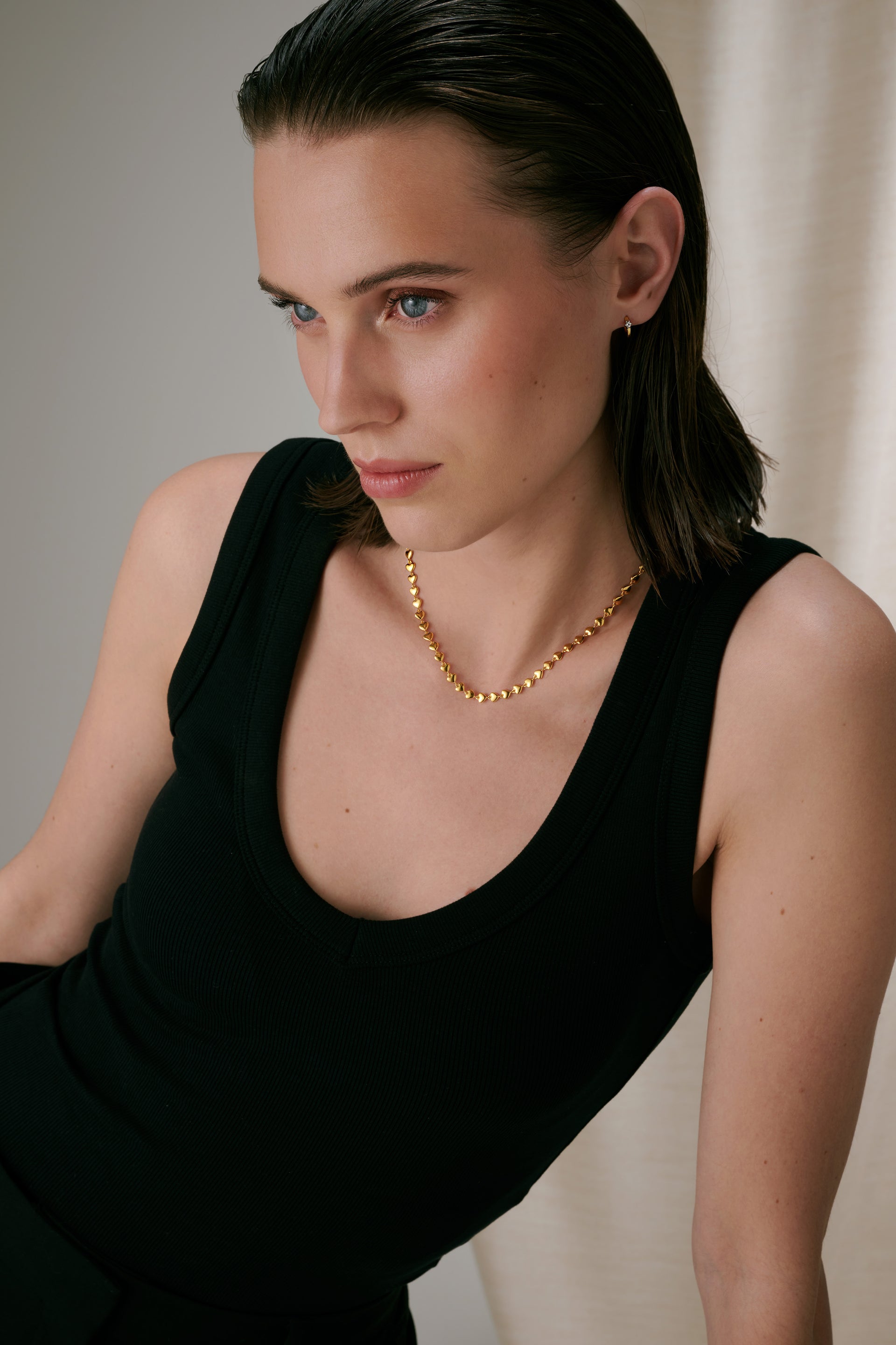 A gold necklace featuring a chain of small connected hearts, paired with a small gold hoop earring.