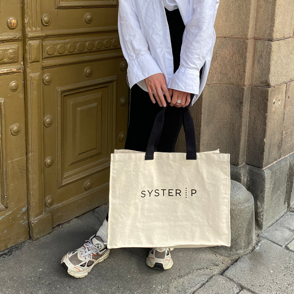 A street style image featuring a model holding the beige Syster P Tote Bag with black handles, while also wearing several silver rings.
