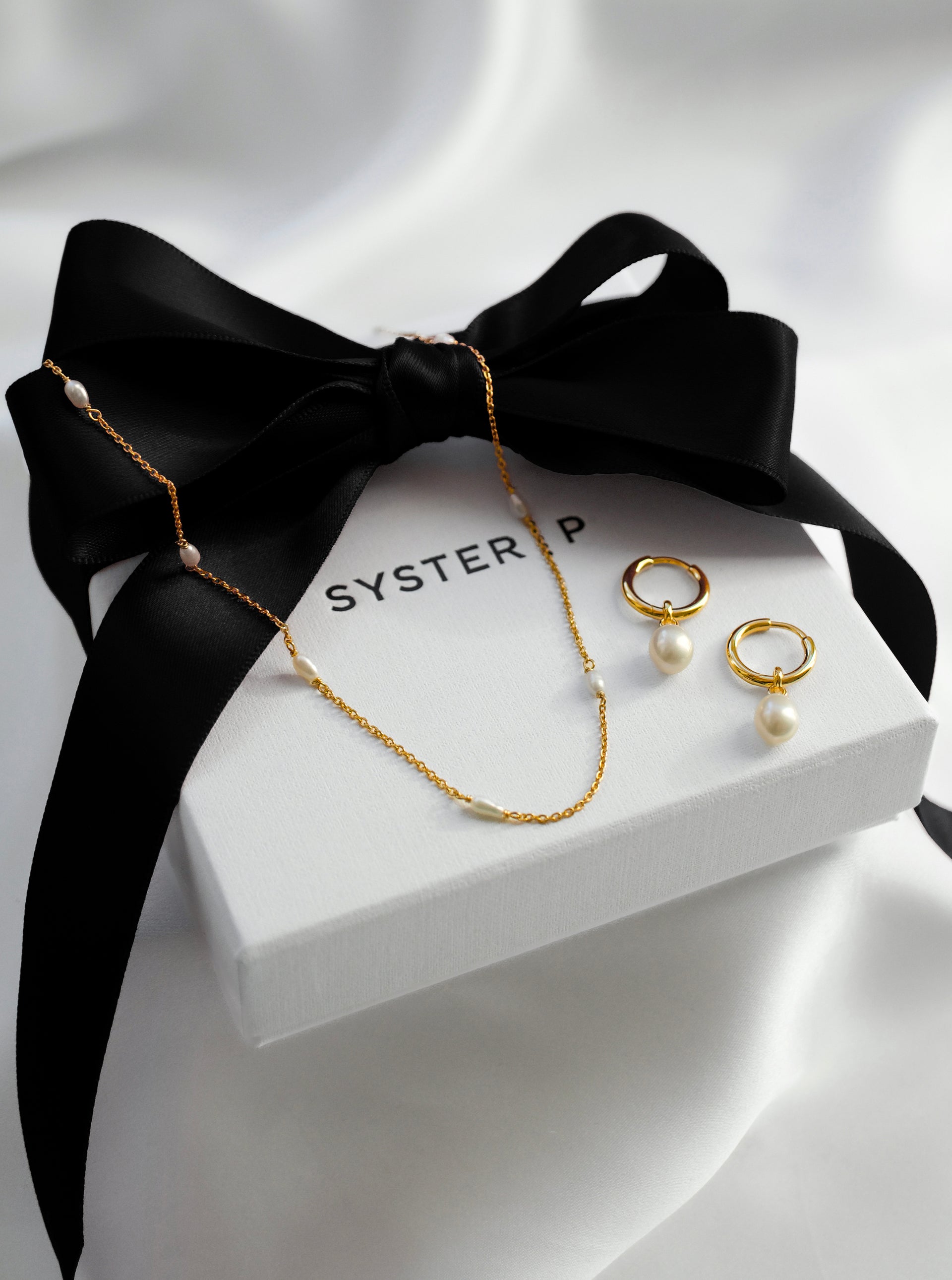 A styled product shot of the gold Treasure Pearl Hoops and the Treasure Multi Pearl Necklace, elegantly arranged on a white Syster P gift box tied with a black ribbon.