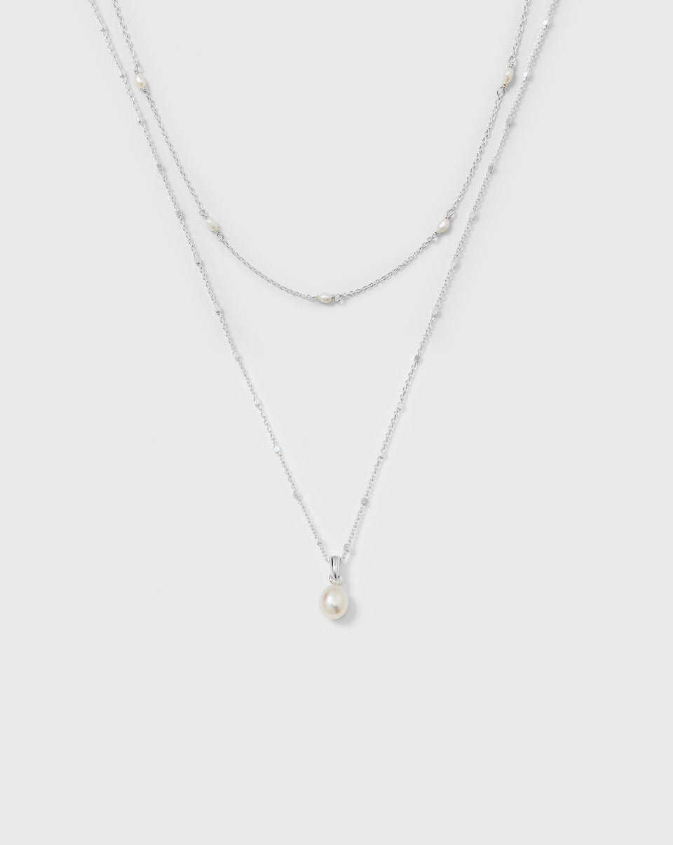 A product shot of the Treasure Necklace Set in silver, featuring two layered necklaces. One necklace has a single pearl pendant, and the other is a delicate chain adorned with multiple small pearls.