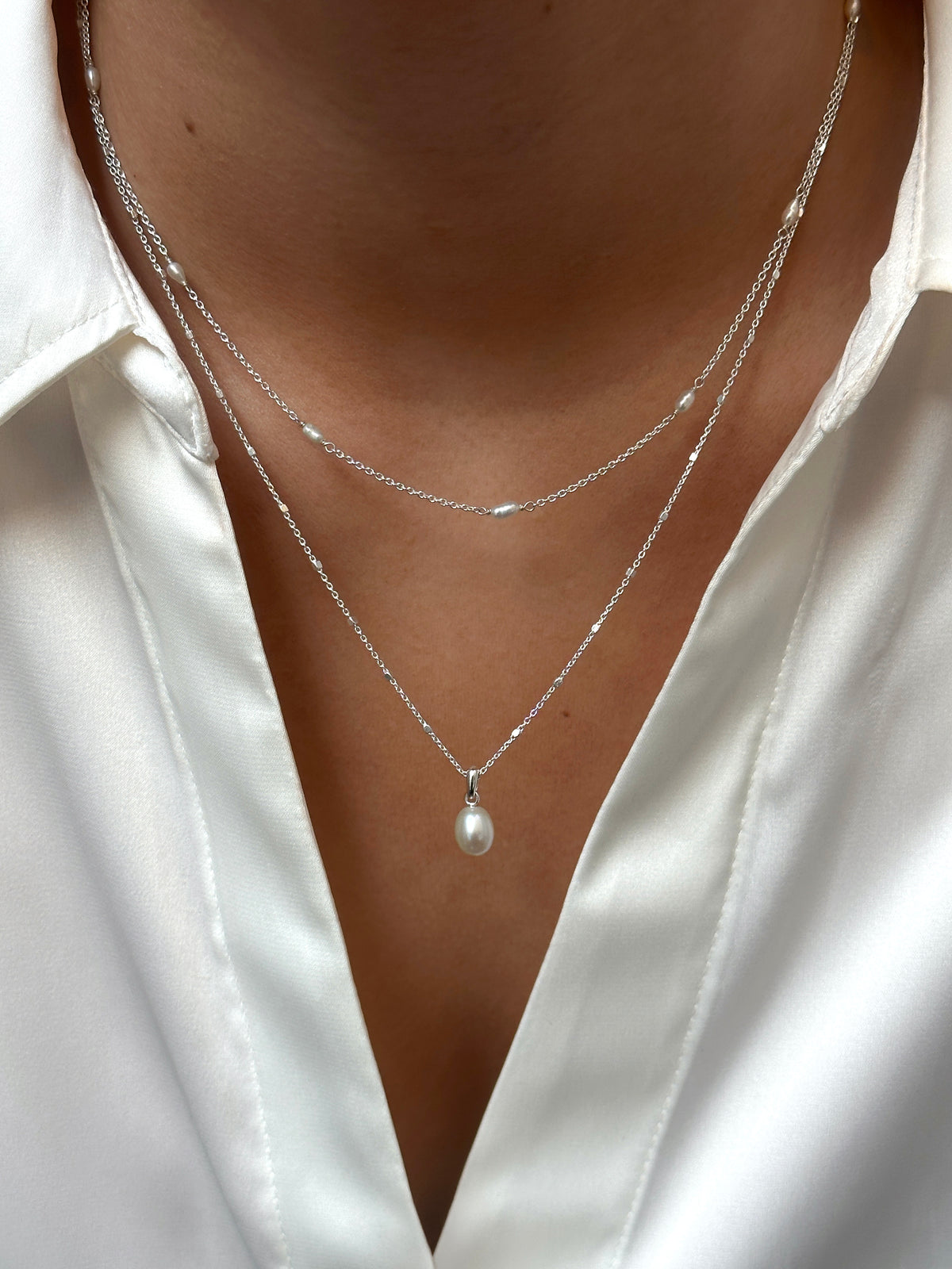 A model wears two layered silver necklaces from the Treasure collection against a white shirt: the Multi Pearl Necklace with small, spaced-out pearls, and the Single Pearl Necklace with a single pearl pendant.