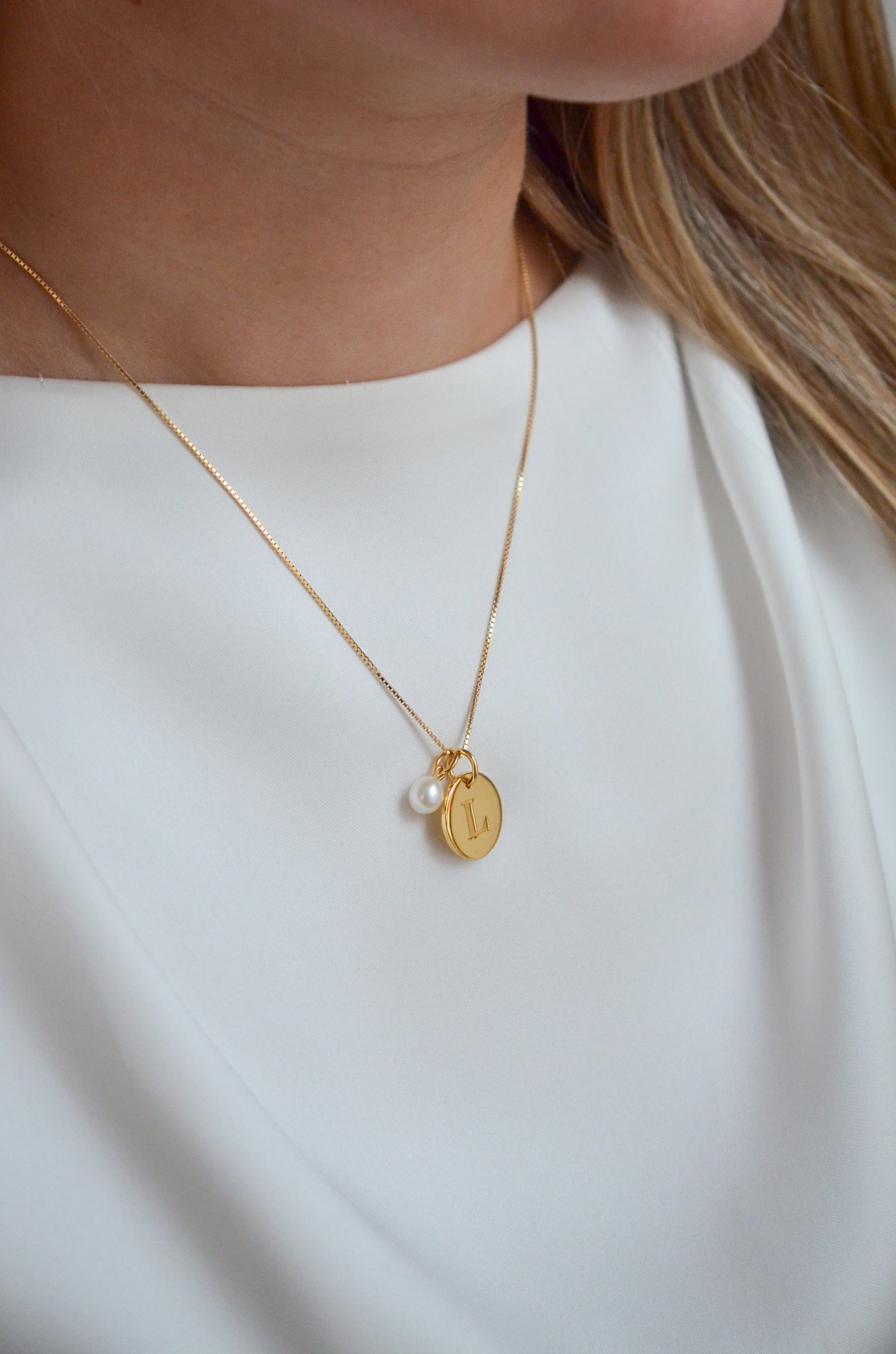 A close-up of a model wearing a gold necklace featuring the Beloved Pearl Pendant alongside a round gold pendant engraved with the letter 'L'.